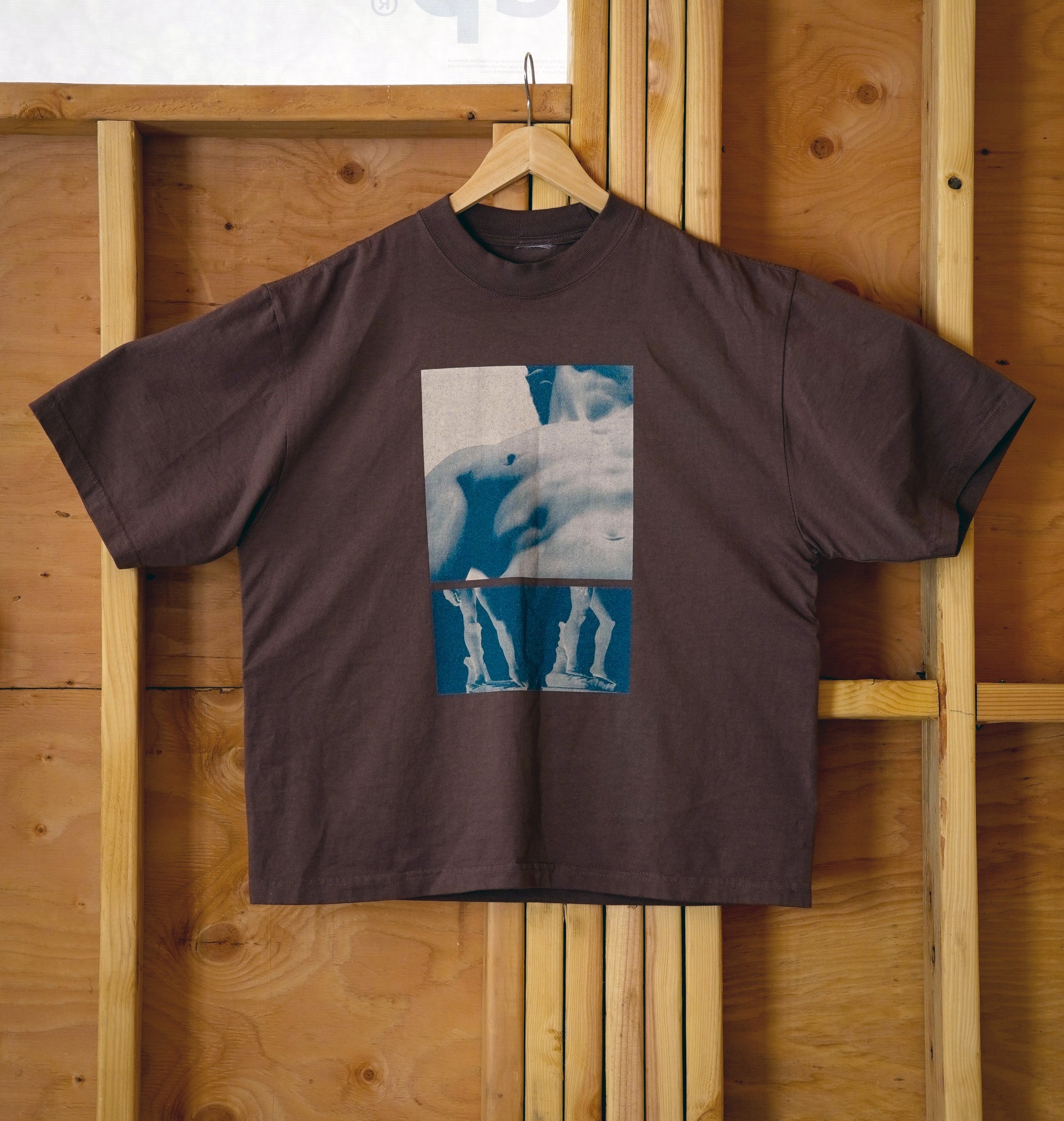 Brown t-shirt with a graphic design on a wooden hanger against a wooden wall.